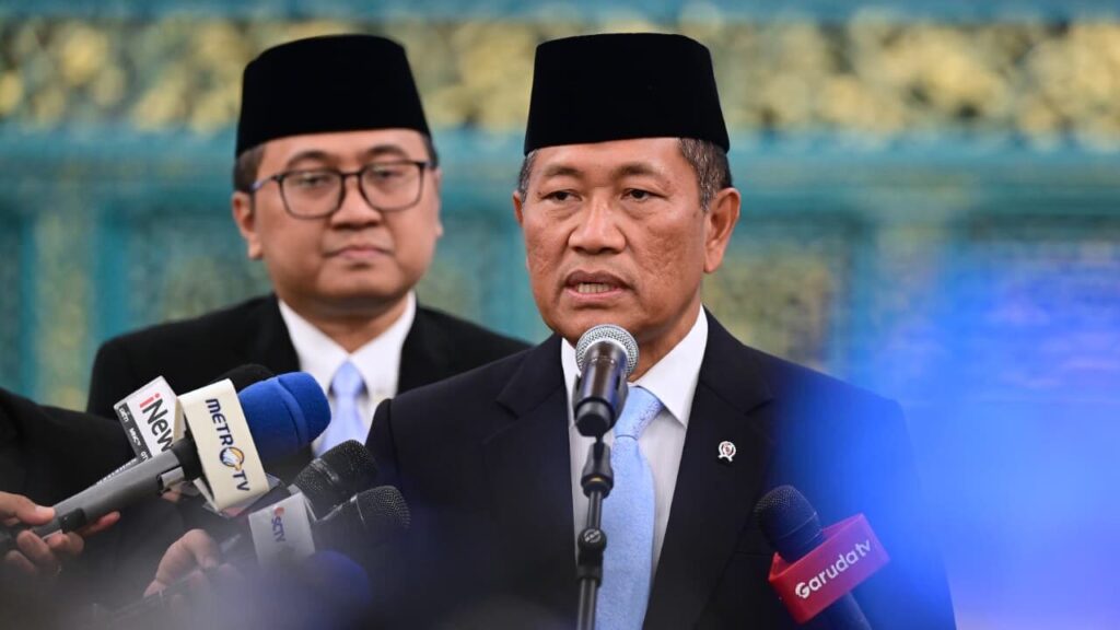 Man in a suit speaks at a microphone during a press conference with reporters and a blurred backdrop behind him